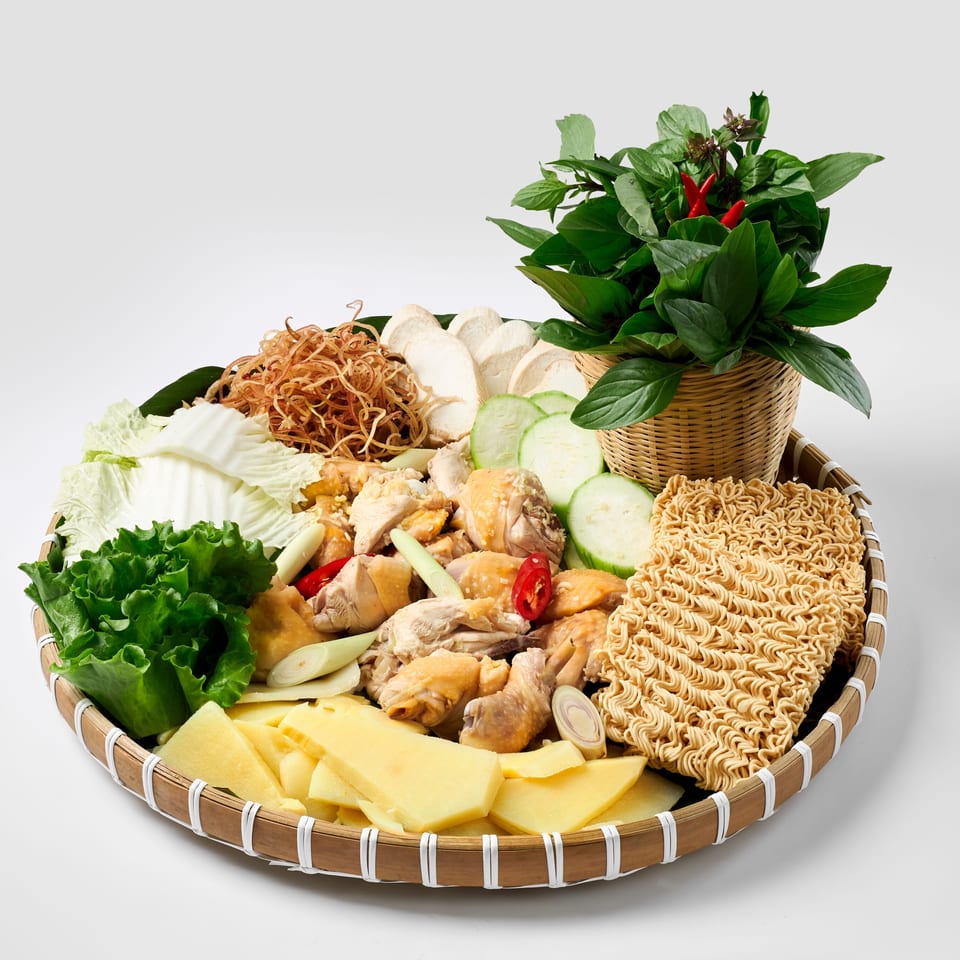 Bamboo tray displaying chicken, instant noodles, vegetables, herbs, chilies, lettuce, cabbage, cucumber, and sliced bamboo shoots.
