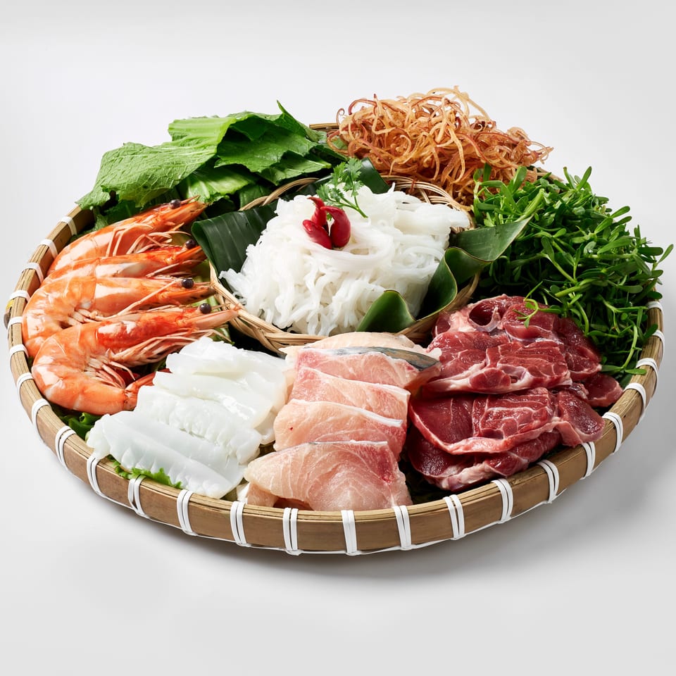Bamboo tray filled with shrimp, sliced meats, fish, noodles, leafy greens, herbs, and vegetables for a Vietnamese hot pot.
