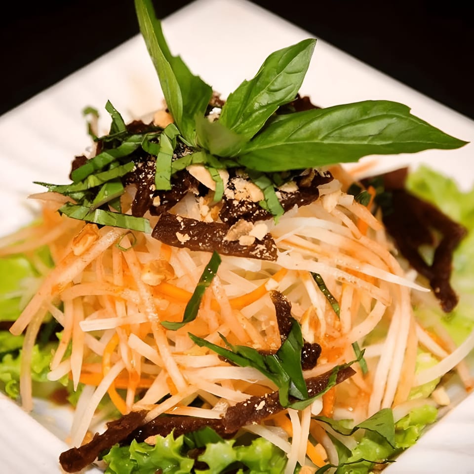 Vietnamese papaya salad with shredded vegetables, dark strips, peanuts, and fresh basil on a white plate.